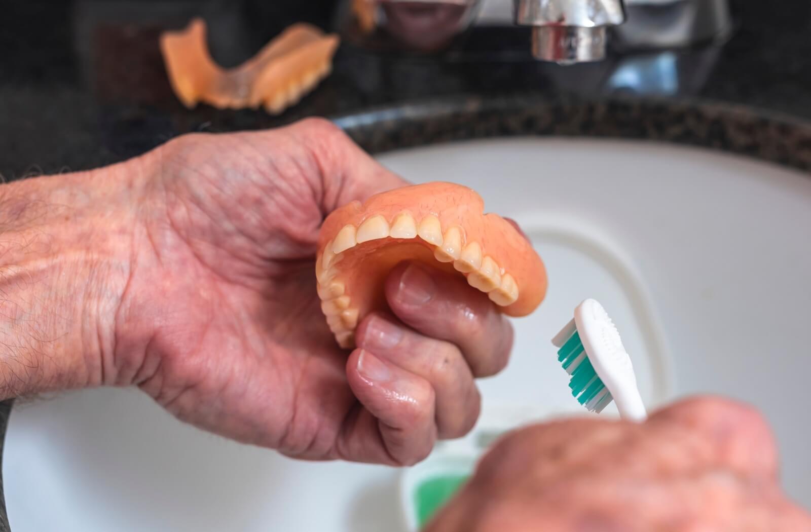A person uses a soft brush to clean and rinse their dentures under a tap.