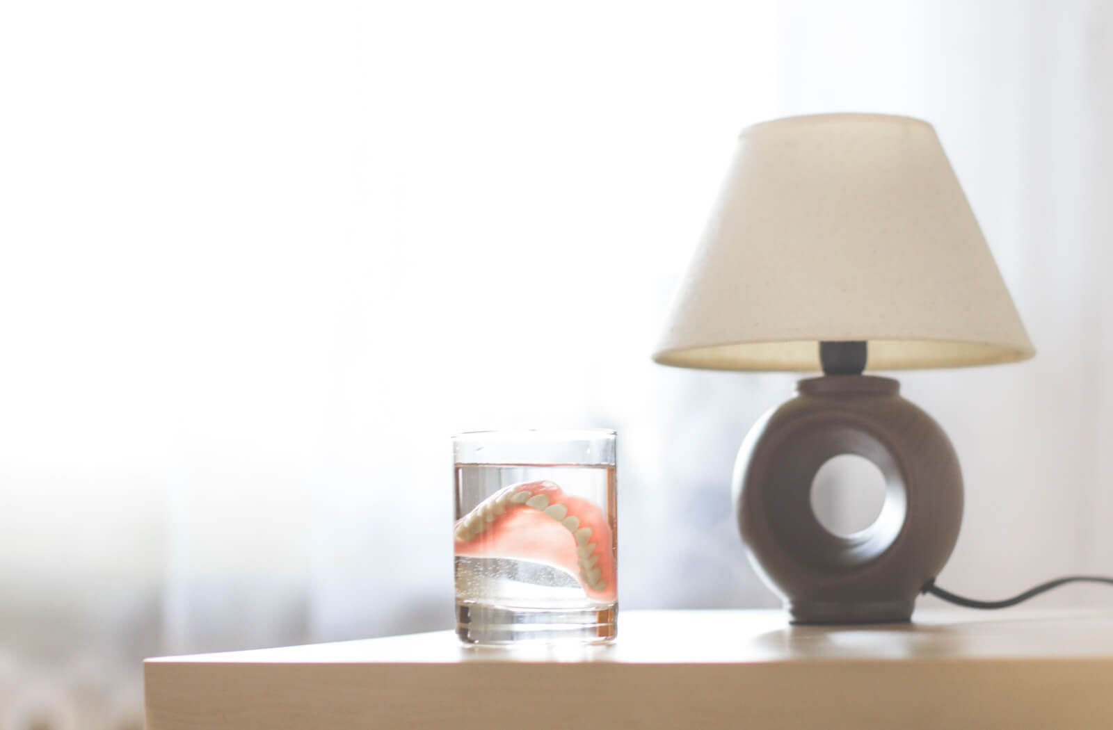 An acrylic set of dentures soaks in a glass of water on a bedside table.