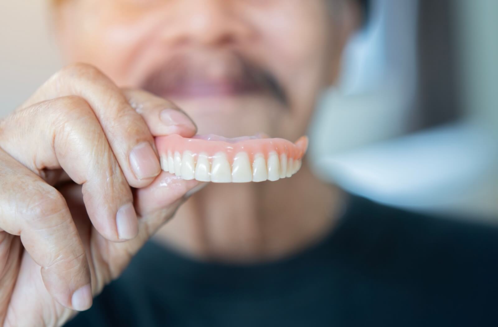 A person holding a set of upper dentures close to the camera with a blurred smiling face in the background