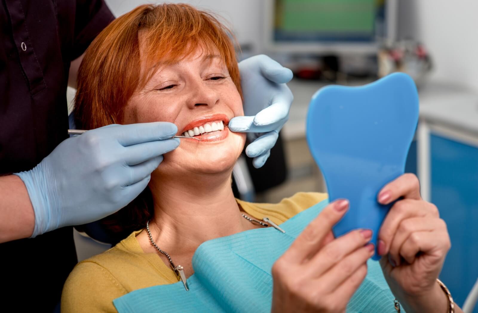 A person sits in a dental chair smiling while holding a mirror, as a dental professional wearing gloves adjusts their dentures during a fitting.